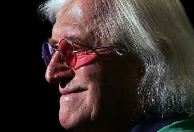 Close-up profile photo of Jimmy Savile in old age wearing round sunglasses with different coloured lenses