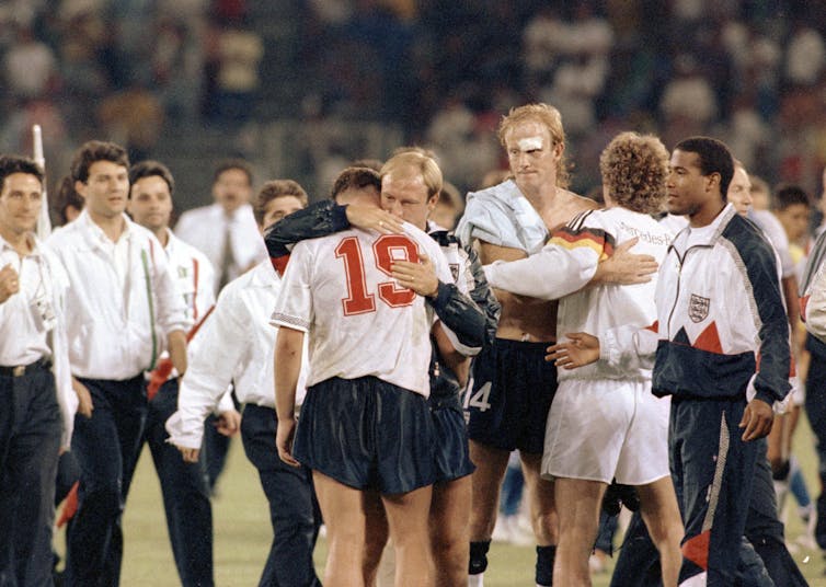 Footballers crowd around on a pitch consoling one wearing a 19 shirt who is crying.
