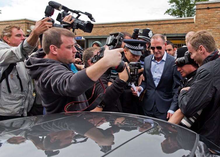 Photographers crowd a man in a blue suit and sunglasses walking towards a car.