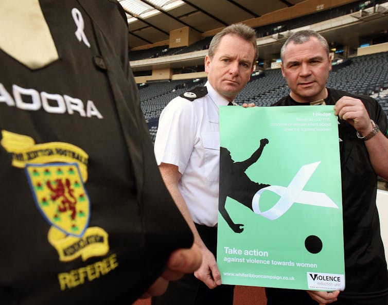 Two men hold up a green poster featuring a football, a white ribbon and a person.