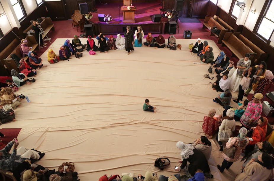 Muslim women, many with heads covered, sit in a large circle, in the center of which is a young boy.