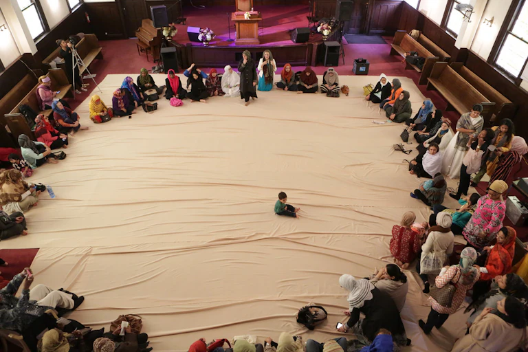 Muslim women, many with heads covered, sit in a large circle, in the center of which is a young boy.