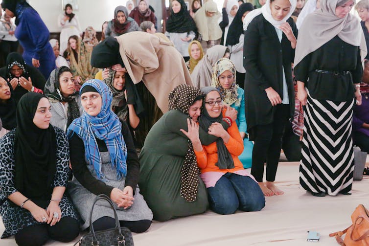 Muslim women in brightly colored clothing and headress kneeling on a rug in a mosque.