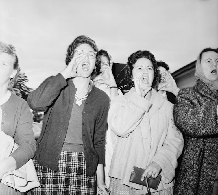 A group of white women are standing across the street from a school and screaming at three black girls.