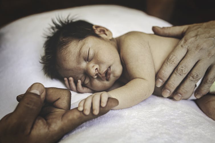 Sleeping baby holding finger of parent with another parent's hand wrapped around their torso