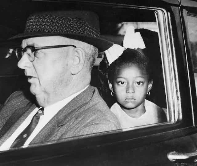 A young black girl is seen in a car sitting behind an older white man. 