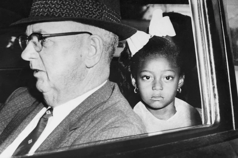 A young black girl is seen in a car sitting behind an older white man.