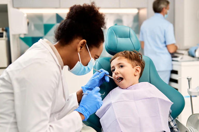 A child in a chair and a woman next to him in a white coat, mask and gloves holding an instrument near his mouth
