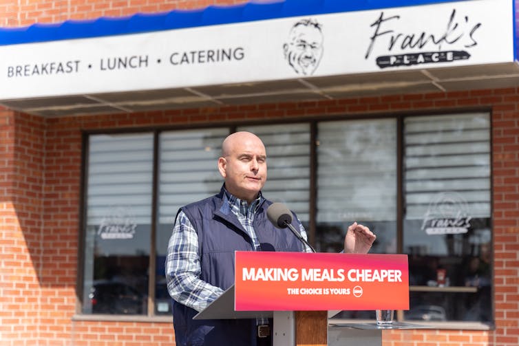 A bald man speaks into a microphone at a podium with a sign that reads Making Meals Cheaper in front of a restaurant called Frank's.