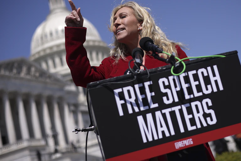 A white woman stand outside the U.S. Capital holding a sign that reads 'Free Speech Matters'