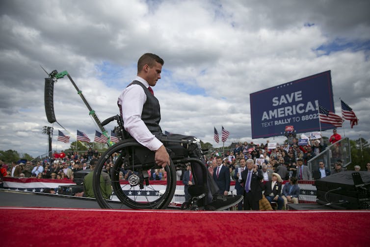 A white man in a wheelchair is on stage during a rally before hundreds of people.