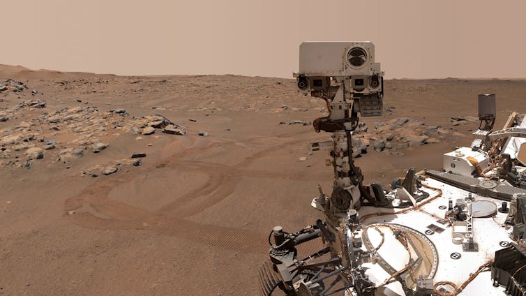 A rust-colored rocky landscape surrounds NASA's Perseverance Mars rover as it rests on Martian soil.