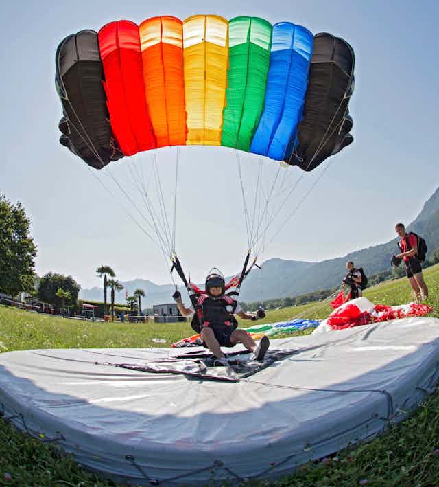 Skydiver lands on a soft white pad while wearing a parachute that is fully deployed and has the colors of the rainbow with black strips at both ends in a large field