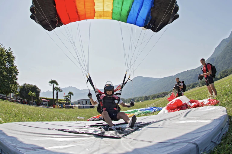 Skydiver lands on a soft white pad while wearing a parachute that is fully deployed and has the colors of the rainbow with black strips at both ends in a large field