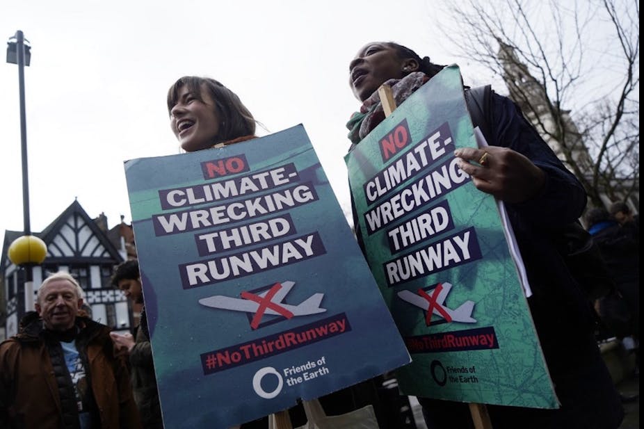 Deux femmes tenant de grands panneaux, avec le texte "non climate-wrecking third runway" et le pictogramme d'un avion en décollage rayé.