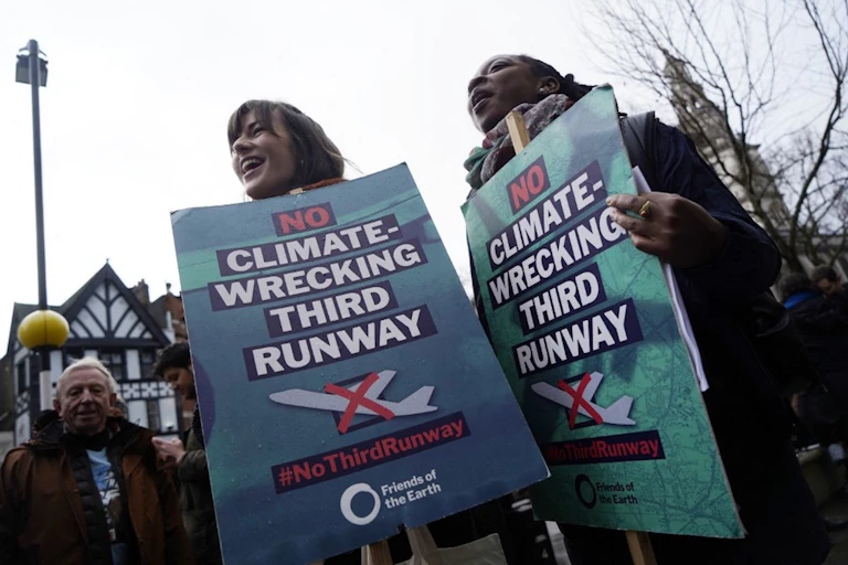 Deux femmes tenant de grands panneaux, avec le texte "non climate-wrecking third runway" et le pictogramme d'un avion en décollage rayé.