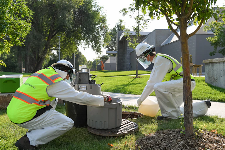 two people in hazmat suits kneel near open manhole