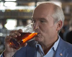 A bald man in a blue jacket and shirt sips on a beer.