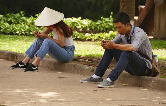 Two people, on wearing a traditional asian conical sunhat, sit on a curb looking at their smartphones.