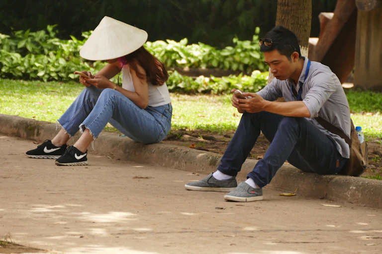 Two people, on wearing a traditional asian conical sunhat, sit on a curb looking at their smartphones.