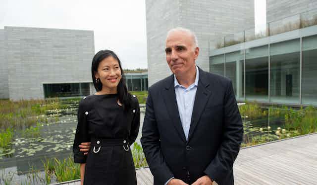 A woman and a man stand on a boardwalk next to a water garden and a modern building.