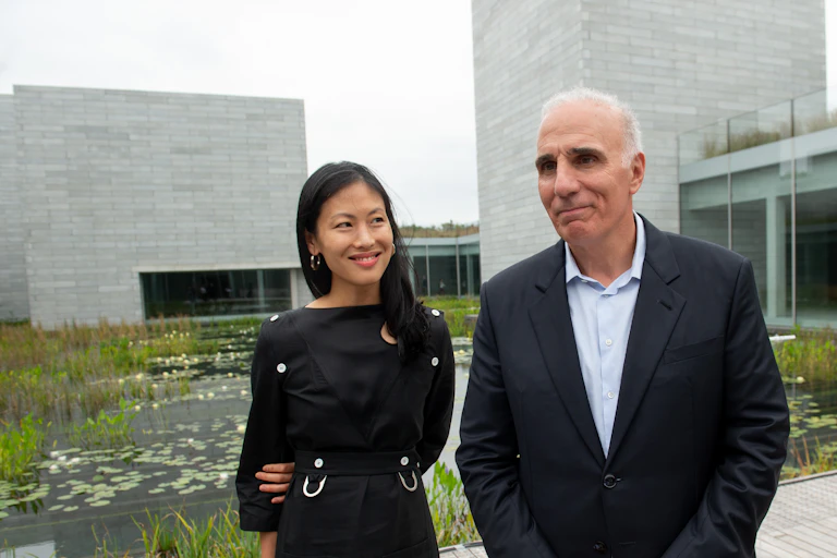 A woman and a man stand on a boardwalk next to a water garden and a modern building.