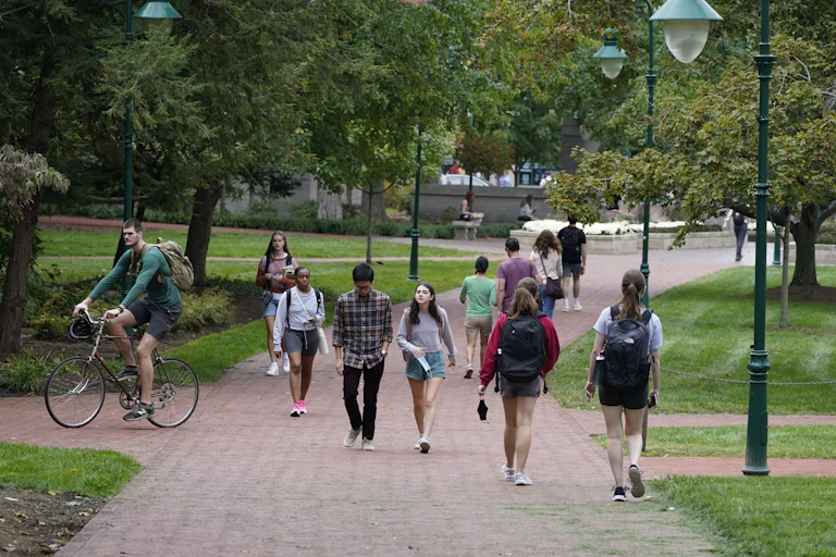 A dozen students stroll across a pathway on a college campus.