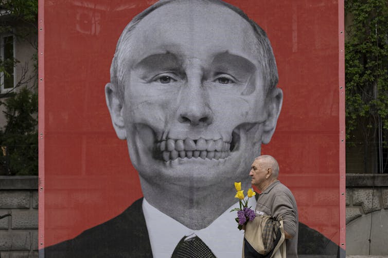 A large mural is seen of a man in a suit depicted as having skeleton teeth and a man walks in front on the street holding flowers.