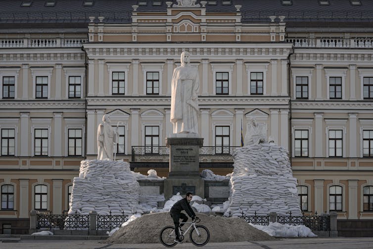 A man seen riding a bicycle past a statue surrounded by sandbags.