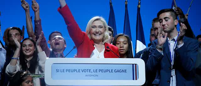 Marine Le Pen, smiling and in a red jacket, waves as a bunch of young-looking supporters cheer.