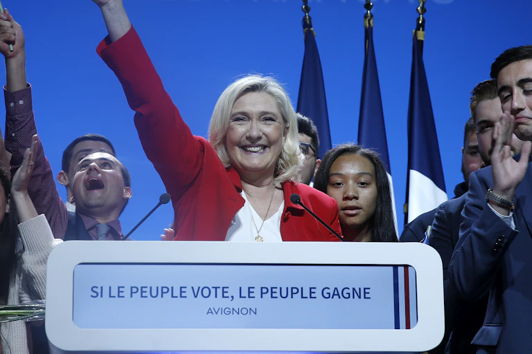 Marine Le Pen, smiling and in a red jacket, waves as a bunch of young-looking supporters cheer.