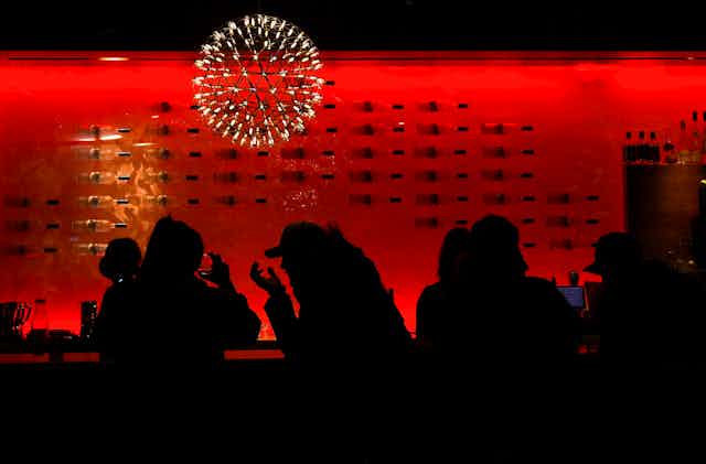 People are silhouetted against a red backdrop and under a chandelier as they sit in a bar. 