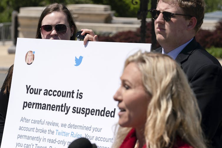 a woman speaks into a microphone and foreground as a man and a woman behind her holdup a poster displaying a message about a Twitter account suspension