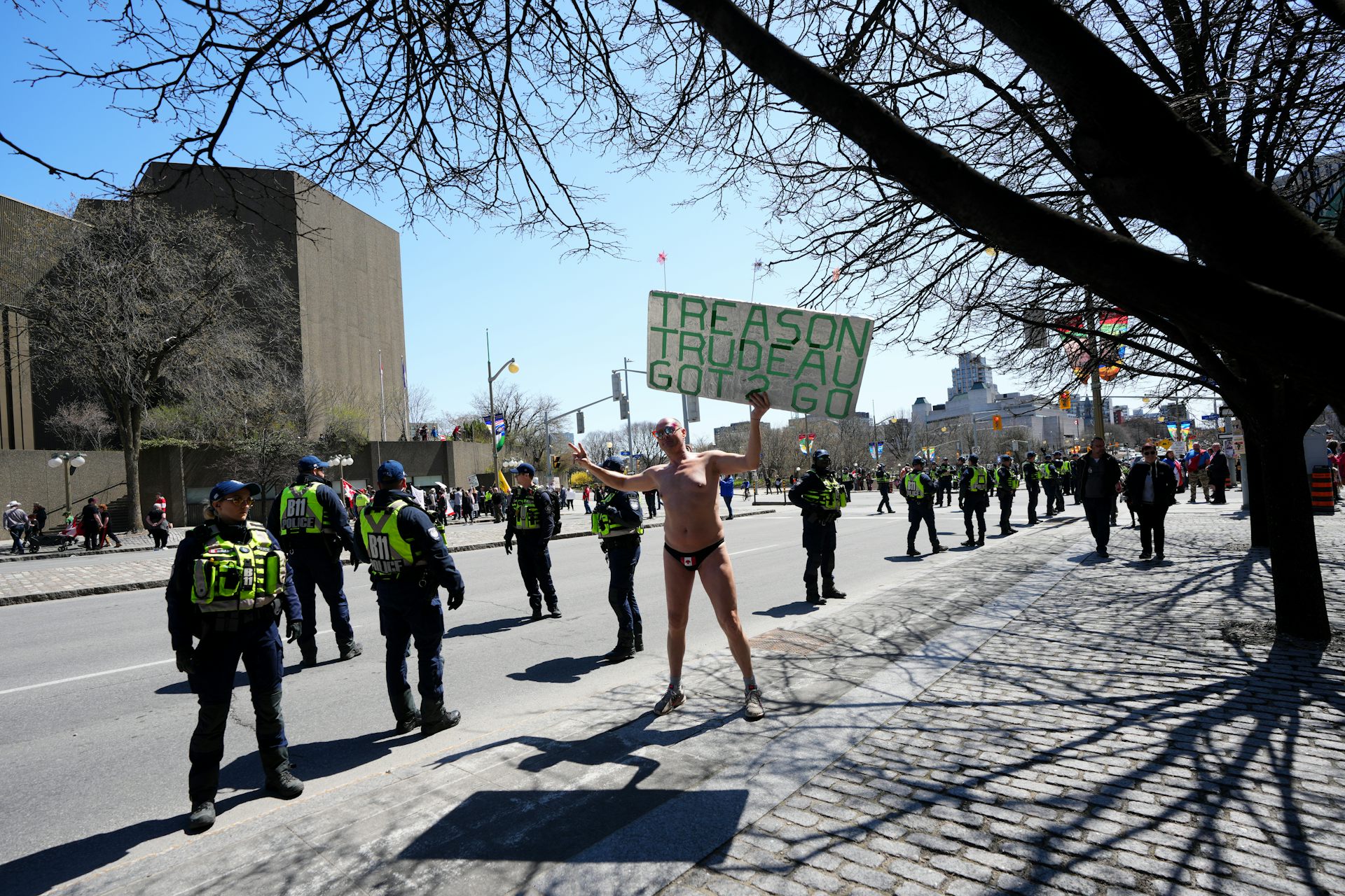 A man naked except for his underwear and shoes holds a sign that reads trudeau treason got to go