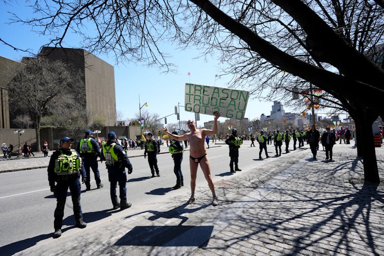 A man naked except for his underwear and shoes holds a sign that reads trudeau treason got to go