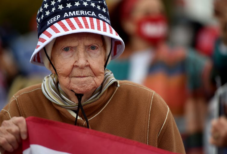 An elderly woman wears a 'Keep America Great Again' hat and holds an American flag.