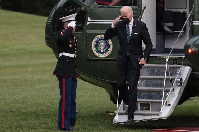 US president, Joe Biden, disembarks from  his helicopter, Marine One, and is saluted by a marine soldier.