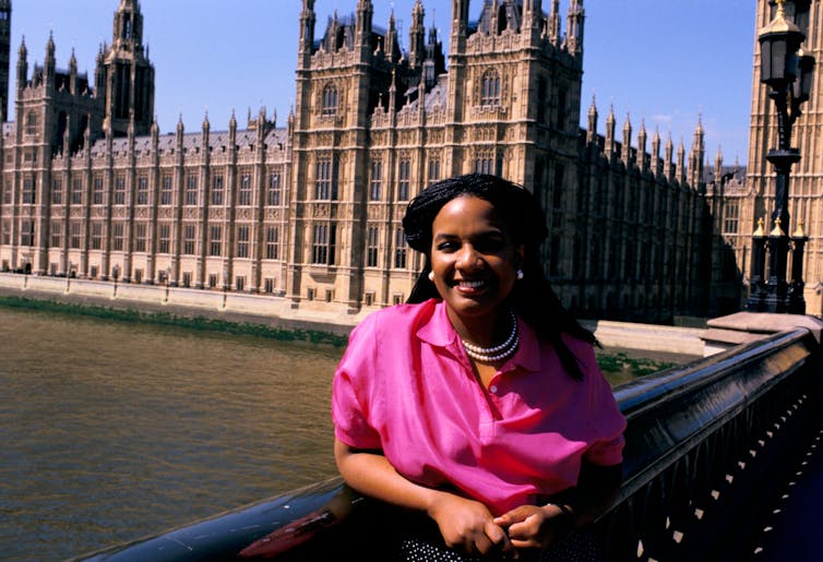 A black woman in pearls and a bright pink shirt stands in front of the houses of parliament.