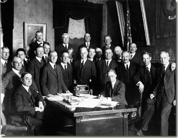 A black and white photo shows men in suits gathered around a desk covered with papers