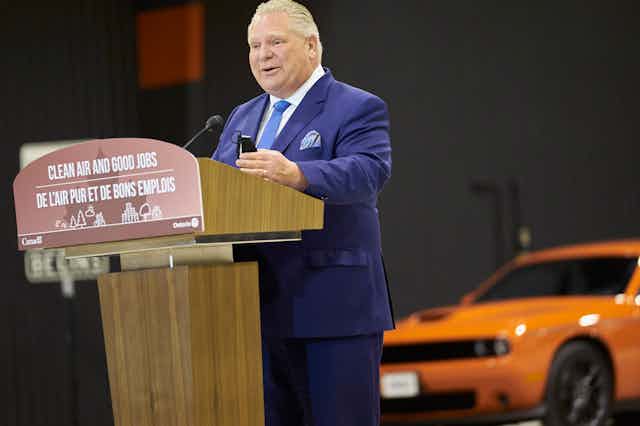 A man in a blue suit stands at a podium with an orange car in the background