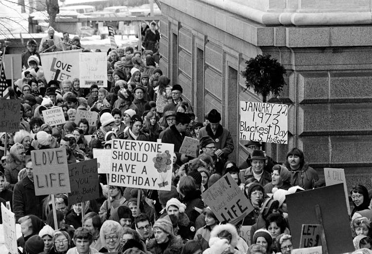 A crowd of men and women protesting near a city building in the winter, holdings signs that say 'Love life' and 'Everyone should have a birthday.'