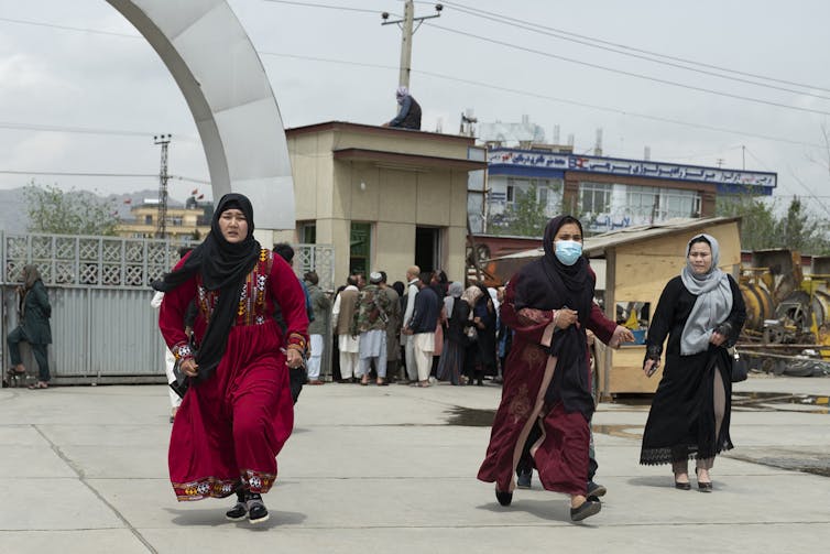 Three Women Wearing Headscarves And Long Dresses Run, Looking Worried, Behind A Group Of Afghan Men