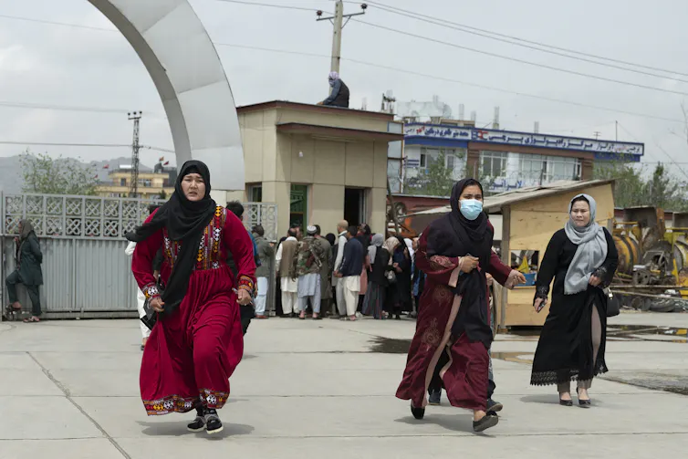 Three women wearing headscarves and long dresses run, looking worried, behind a group of Afghan men