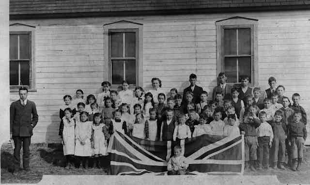 Black and white photo showing a 1908 scene of a schoolteacher in a tie and jacket and white schoolchildren behind a union jack flag 