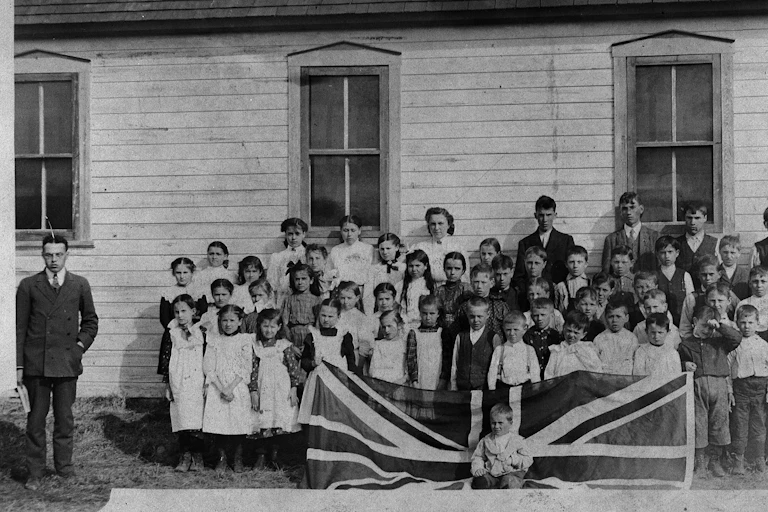 Black and white photo showing a 1908 scene of a schoolteacher in a tie and jacket and white schoolchildren behind a union jack flag