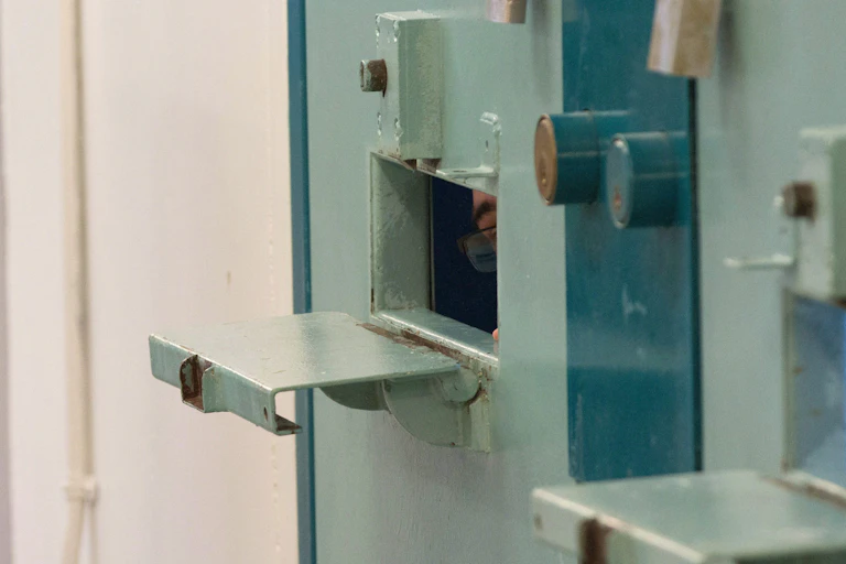 A sliver of the face of an inmate is seen inside a tiny slot in a prison cell door.