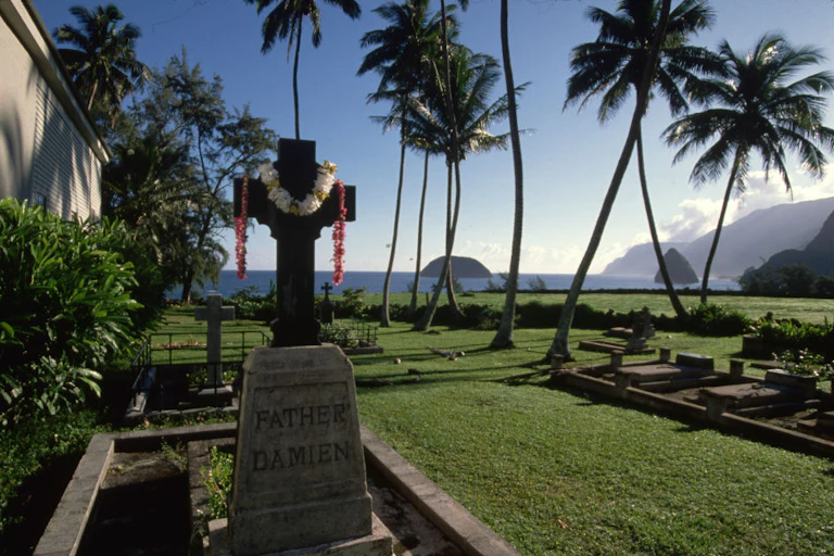 A stone cross monument marks a grave close to the ocean and palm trees.