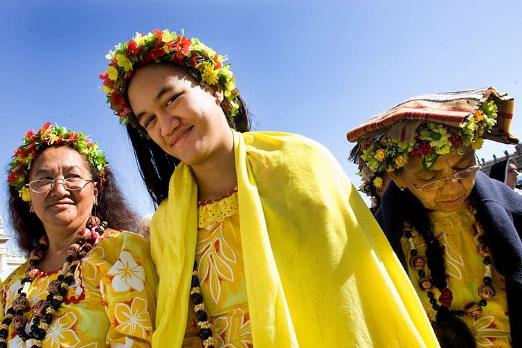 Three women wearing flowers in their hair and dressed in yellow smile at the camera.