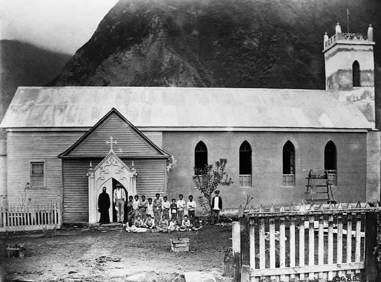 A black and white photograph shows a small group gathered in front of a church in front of a misty mountain.