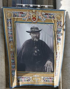 A tapestry with a colored border depicts a portrait of Father Damien, wearing a hat and glasses.